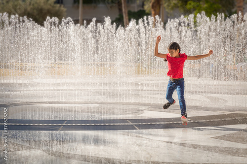She jumps over the water jets just before they shoot up a layered wall of water. A courageous young girl plays running in and out of the modern water fountain.
