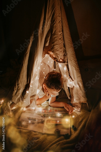magic hut with garlands and lights for the child do it yourself from improvised means. blondy preschooler boy reads a book in toy hut at home