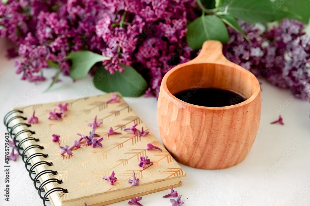 Wooden finnish cup with coffee on the table. Nearby lies a closed notebook for notes and a bouquet of lilacs. Good morning and good mood, planning a day at breakfast, concept.