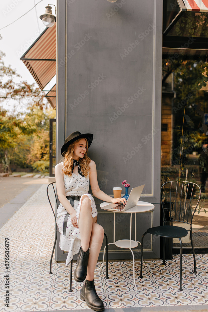 Full-length vertical photo of attractive blonde wearing a hat. Happy girl in trendy summer clothes sitting at laptop in European cafe Full-length vertical photo of attractive blonde wearing a hat. Happy girl in trendy summer clothes sitting at laptop in European cafe