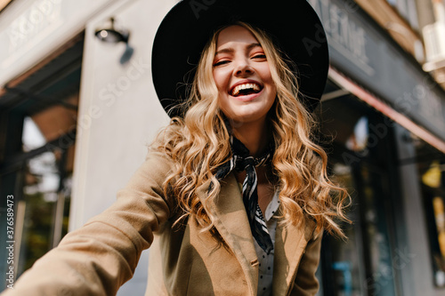 Cute blonde in a hat and a fashionable scarf around her neck makes a selfie. Portrait of a girl with burning cheerful eyes and an amazing smile.