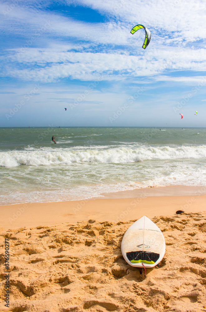 Obraz premium Surfboard on a sand beach. Kitesurfers in the sea in the background. Blue sky with light clouds