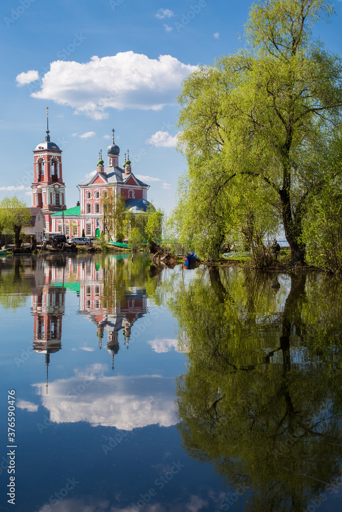 Russian orthodox church, big birch tree, blue sky with clouds reflect in the still river.