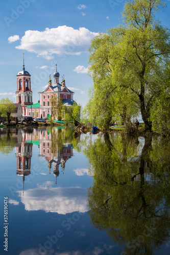 Russian orthodox church, big birch tree, blue sky with clouds reflect in the still river.