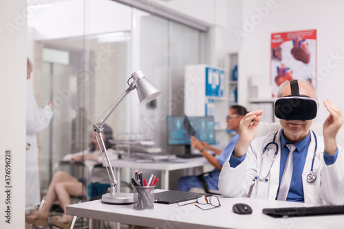 Elderly aged doctor using virtual reality goggles in hospital office. Young therapist talking with disabled woman in wheelchair on clinic corridor and nurse is holding x-ray.