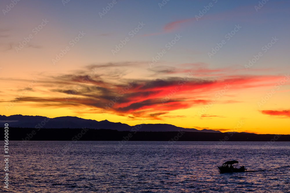 The clouds reflect the colorful sunset over Puget Sound as the sun slowly sets behind the mountains