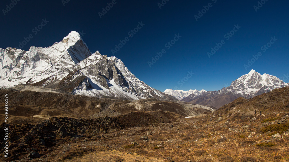 Snow-capped Mt Ama Dablam panorama in the Himalayas. View through a valley with a glacier on the left and a trekking path on the right. Clear bright blue sky.