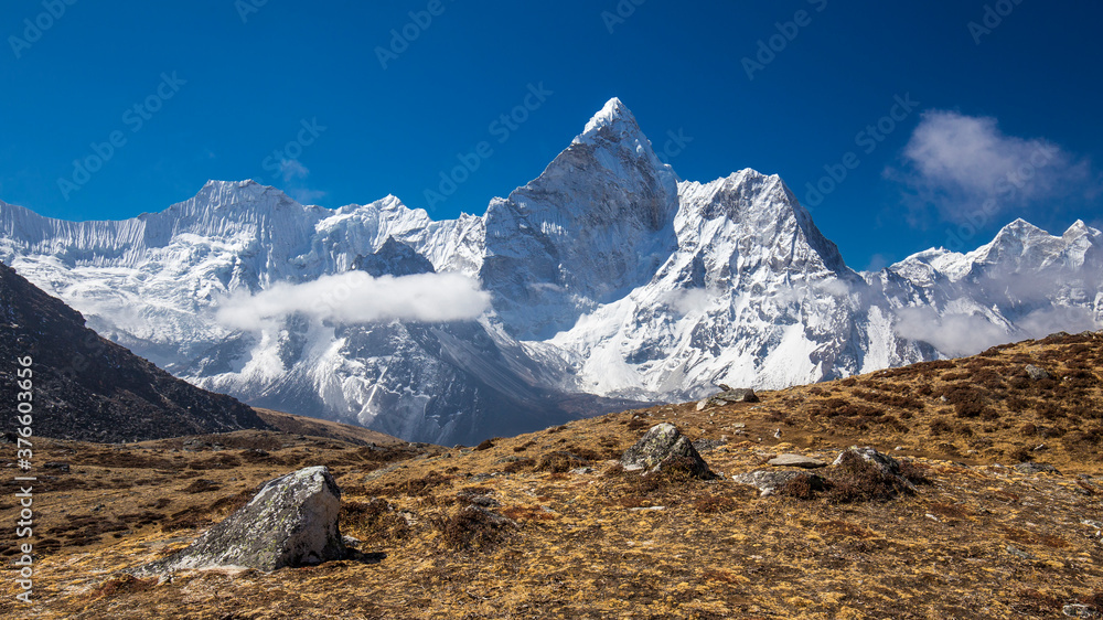Snow-capped Mt Ama Dablam panorama in the Himalayas. View from the yellowish top of a hill along a trekking path. Bright blue sky with light clouds. 