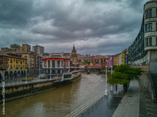 Cityscape of Bilbao, Spain, with the market hall and the spire of the church San Anton aside the river Nervion with colorful buildings and a dark cloudy sky
