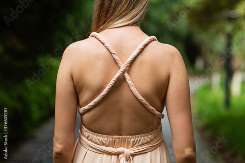 Elegant young woman with blonde hair posing in maxi formal dress from behind outdoor at summer