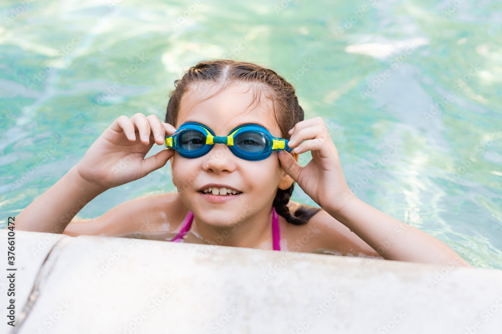 girl in pool touching swim goggles while looking at camera