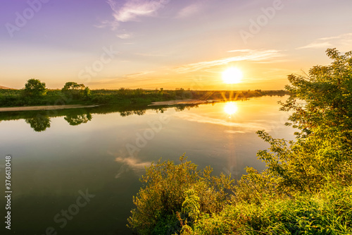 Fototapeta Naklejka Na Ścianę i Meble -  Scenic view at beautiful summer river sunset with reflection on water with green bushes, grass, golden sun rays, calm water ,deep blue cloudy sky and glow on a background, spring evening landscape