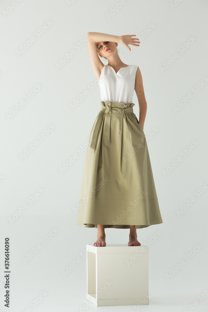 Woman model stands barefoot on a cube in the studio on a light background. Clothes - long skirt and white summer T-shirt. The arm is bent and covers her forehead.