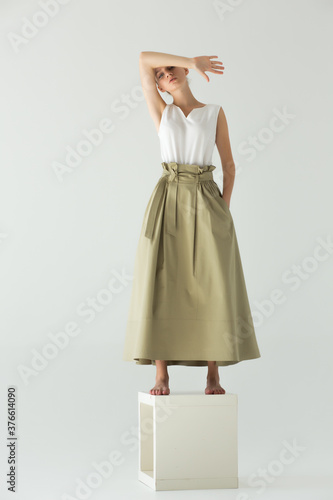 Woman model stands barefoot on a cube in the studio on a light background. Clothes - long skirt and white summer T-shirt. The arm is bent and covers her forehead.