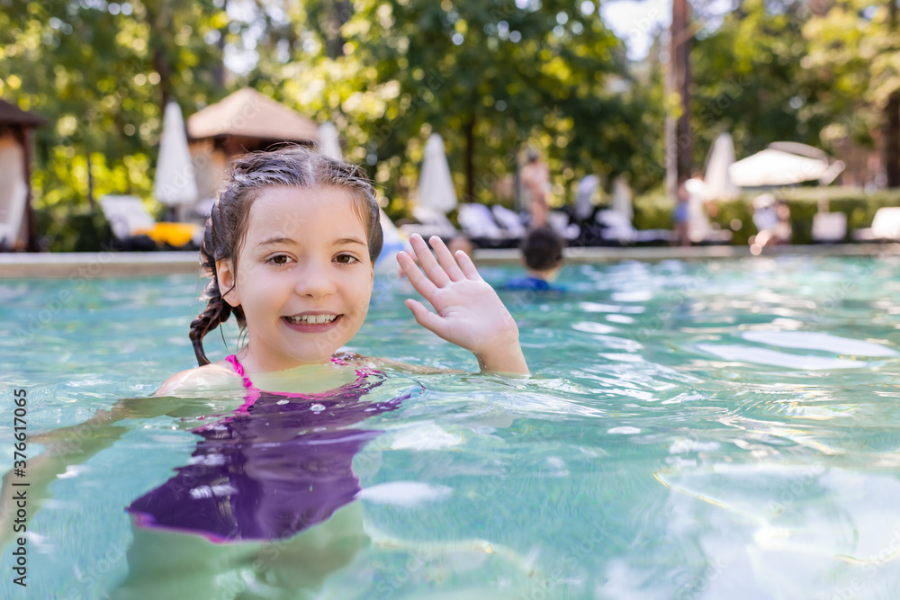 joyful girl in swimming pool looking at camera and waving hand