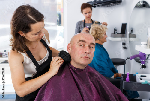Adult bald male client receiving service by professional barber in salon