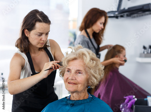 Skilled woman hairdresser making hairstyle for elderly female client in hair studio..