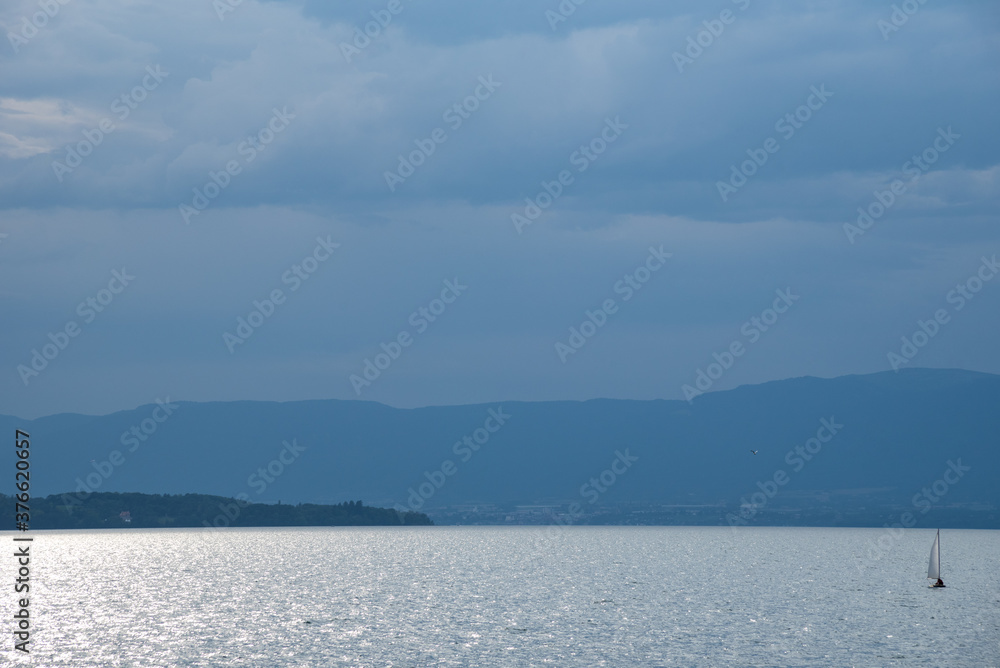 Sailing boat on Geneva Lake at evening with beautiful cloudscape and sun light glow on the calm water. View of Swiss side from French shore. Solitude, relaxing vacation concepts.
