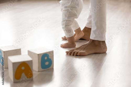 Close up barefoot african american woman teaching little biracial baby kid walking on warm wooden floor indoors. Happy mixed race family involved in daycare activity, making first steps in living room