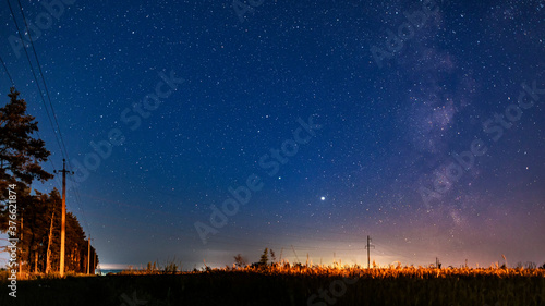The Milky Way, Saturn, Jupiter and many stars in the night sky in a field near the country road and power line. Cosmic landscape.