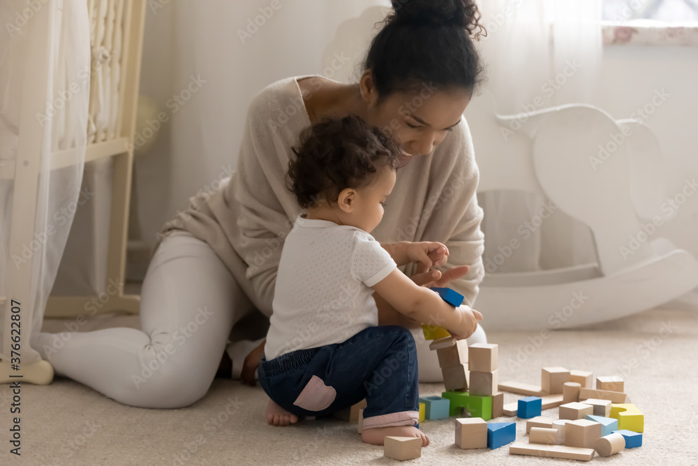 Smiling young mixed race woman babysitter enjoying playing with little adorable funny baby boy or girl in children bedroom, affectionate african mother constructing building with cubes with small kid. Smiling young mixed race woman babysitter enjoying playing with little adorable funny baby boy or girl in children bedroom, affectionate african mother constructing building with cubes with small kid.