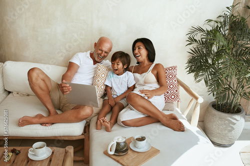 Happy Family On Sofa. Portrait Of Mixed Race Parents With Little Son Using Tablet And Enjoying Leisure On Summer Vacation At Tropical Resort. Different Ethnic Mom And Dad With Boy On Weekend. photo