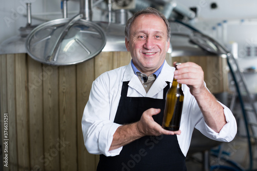 Portrait of brewer who is standing with bottle for beer on his workplace in the brew-house.