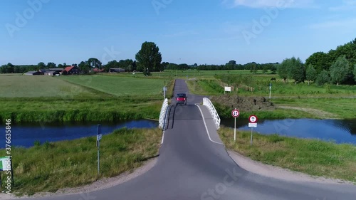 Low altitude aerial view of red microcar driving over small bridge drone moving above road and vehicle scenery polder landscape also showing farm in the background 4k high resolution footage photo