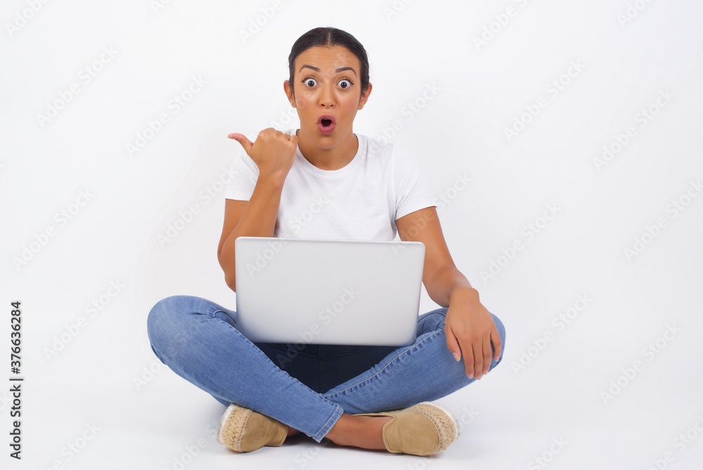 Horizontal shot of pretty young joyful shocked female points with thumb away, indicates something on blank wall isolated over white background. Check this out. Ethnicity concept. Horizontal shot of pretty young joyful shocked female points with thumb away, indicates something on blank wall isolated over white background. Check this out. Ethnicity concept.