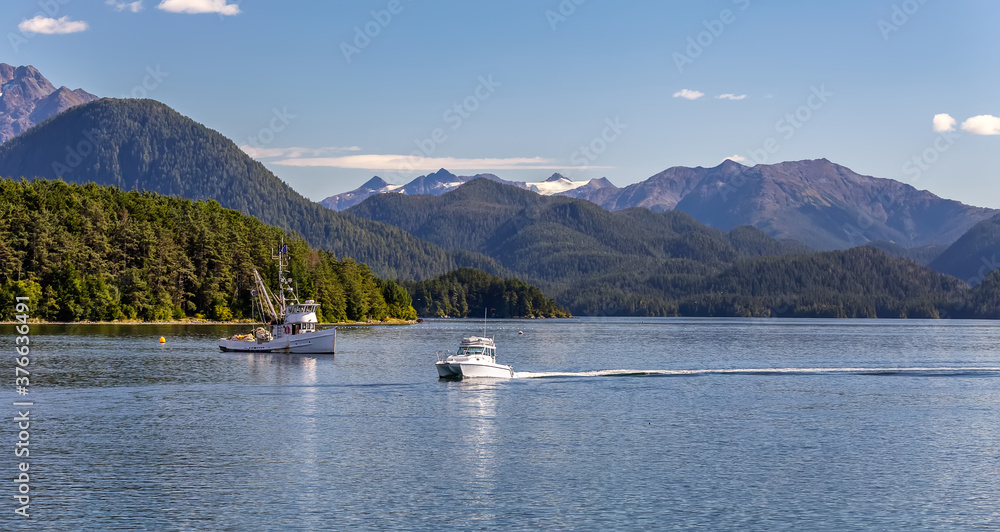A beautiful panoramic shot of a harbour in Sitka, AK with a commercial fishing boat anchored in it and a small speedboat sailing, green forest, mountains with snowy peaks, and gorgeous blue sky.