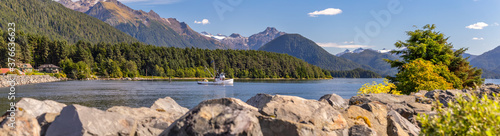 Beautiful panoramic shot of a harbour in Sitka, AK. Commercial fishing boat anchored in it, green forest, mountains with snowy peaks, and gorgeous blue sky. Blurred stone barrier in the foreground.