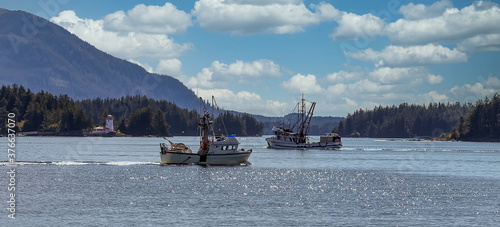 Shot of commercial fishing boats sailing in a harbour in Sitka, AK. Green forest, mountains, beautiful blue sky with clouds, and a lighthouse in the background.