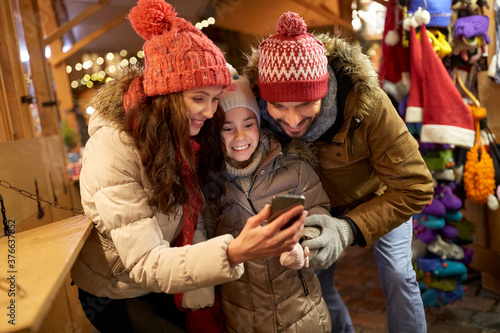 family, winter holidays and technology concept - happy mother, father and little daughter with smartphone at christmas market in evening