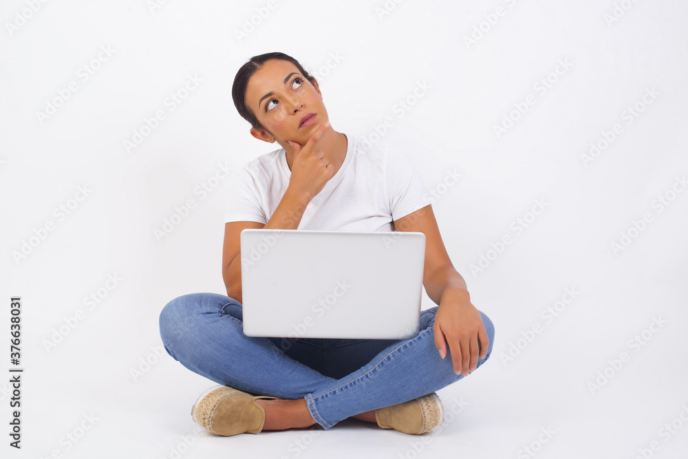 Portrait of thoughtful smiling girl keeps hand under chin, looks sideways, thinking or wondering about something with interest, dressed casually, poses against gray studio. Taking decisions concept. Portrait of thoughtful smiling girl keeps hand under chin, looks sideways, thinking or wondering about something with interest, dressed casually, poses against gray studio. Taking decisions concept.