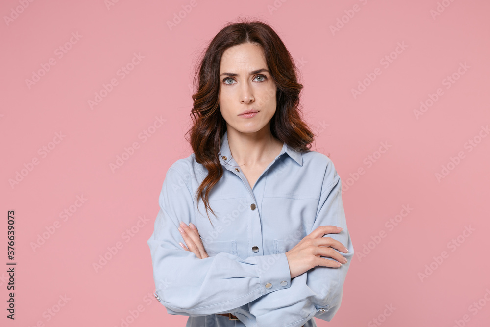 Displeased serious attractive young brunette woman 20s wearing casual blue shirt dress posing standing holding hands crossed looking camera isolated on pastel pink colour background studio portrait. Displeased serious attractive young brunette woman 20s wearing casual blue shirt dress posing standing holding hands crossed looking camera isolated on pastel pink colour background studio portrait.