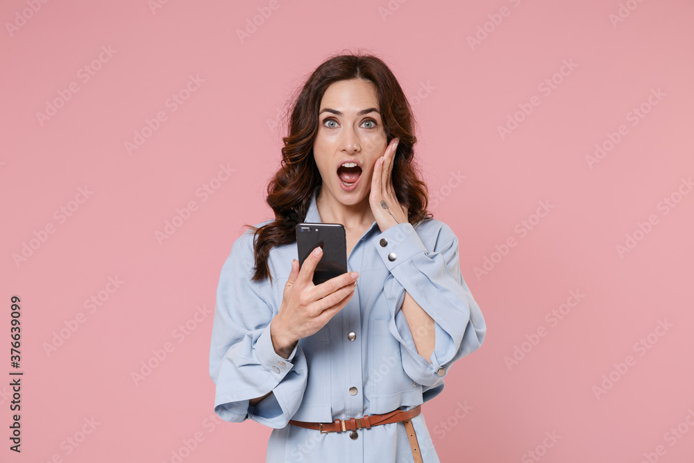 Shocked amazed young brunette woman 20s wearing blue shirt dress using mobile cell phone typing sms message put hand on cheek looking camera isolated on pastel pink colour background, studio portrait. Shocked amazed young brunette woman 20s wearing blue shirt dress using mobile cell phone typing sms message put hand on cheek looking camera isolated on pastel pink colour background, studio portrait.