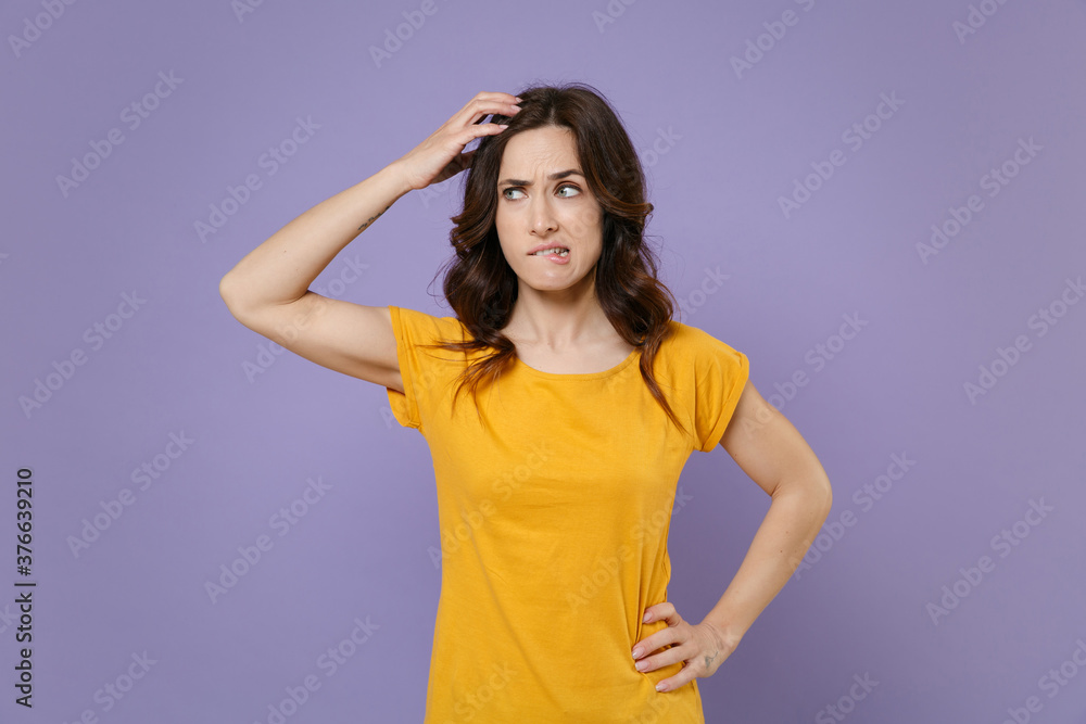 Preoccupied puzzled concerned young brunette woman 20s wearing basic yellow t-shirt posing put hand on head biting lips looking aside isolated on pastel violet colour background, studio portrait. Preoccupied puzzled concerned young brunette woman 20s wearing basic yellow t-shirt posing put hand on head biting lips looking aside isolated on pastel violet colour background, studio portrait.