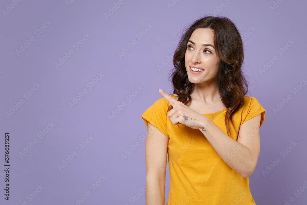 Smiling attractive young brunette woman 20s wearing basic yellow t-shirt posing stand pointing index finger aside up on mock up copy space isolated on pastel violet colour background, studio portrait. Smiling attractive young brunette woman 20s wearing basic yellow t-shirt posing stand pointing index finger aside up on mock up copy space isolated on pastel violet colour background, studio portrait.