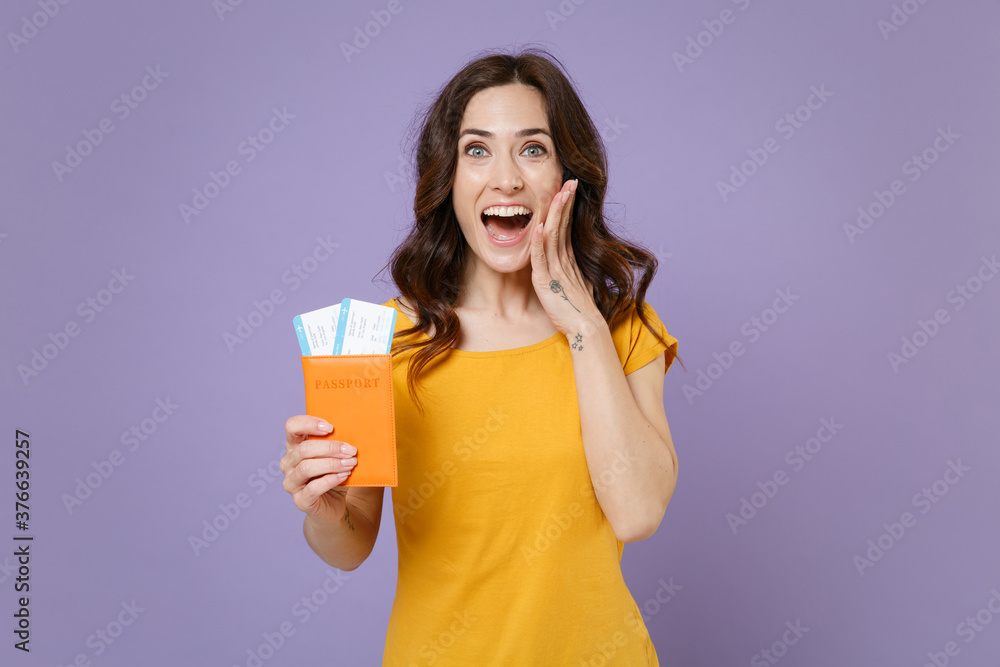 Surprised young brunette traveler tourist woman 20s wearing basic yellow t-shirt hold passport tickets boarding pass put hand on cheek isolated on pastel violet colour background, studio portrait. Surprised young brunette traveler tourist woman 20s wearing basic yellow t-shirt hold passport tickets boarding pass put hand on cheek isolated on pastel violet colour background, studio portrait.
