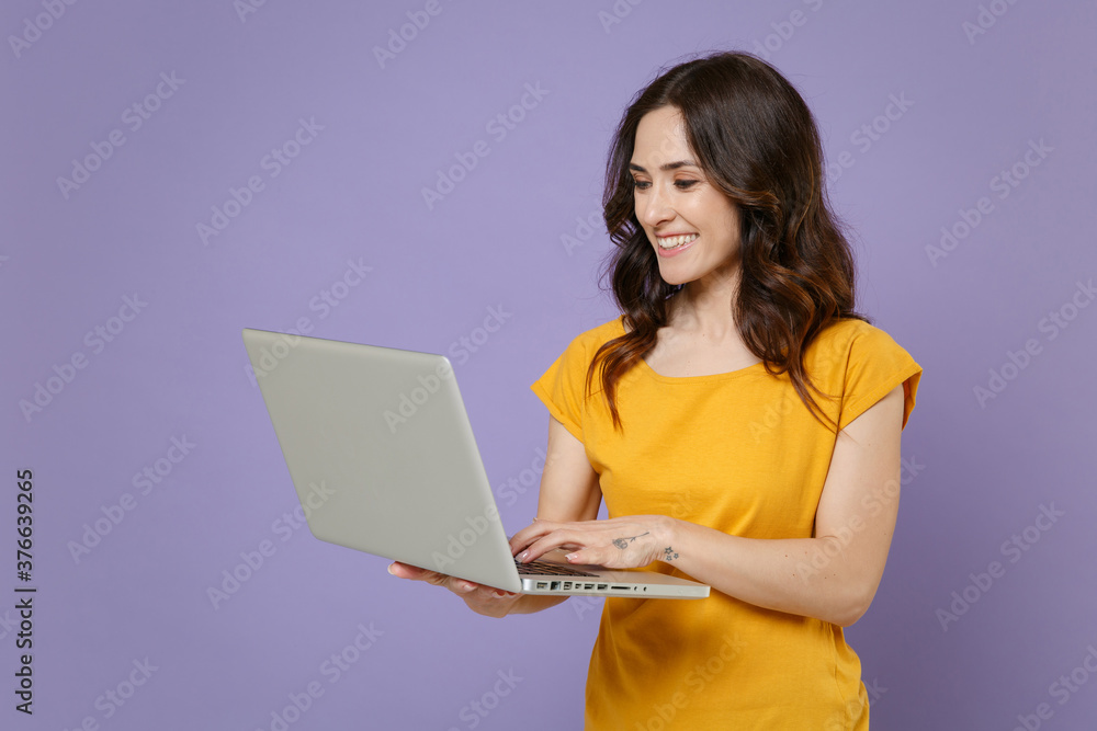 Smiling beautiful pretty young brunette woman 20s wearing basic yellow t-shirt posing standing hold in hands working on laptop pc computer isolated on pastel violet colour background, studio portrait. Smiling beautiful pretty young brunette woman 20s wearing basic yellow t-shirt posing standing hold in hands working on laptop pc computer isolated on pastel violet colour background, studio portrait.