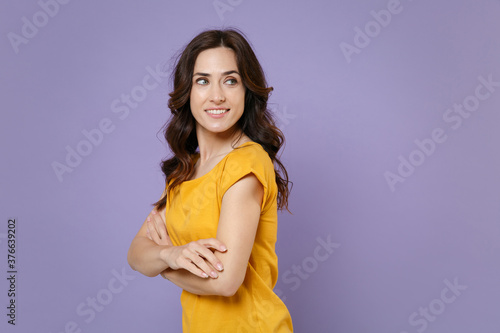 Side view of charming attractive young brunette woman 20s wearing basic yellow t-shirt posing standing holding hands crossed looking aside isolated on pastel violet colour background, studio portrait.