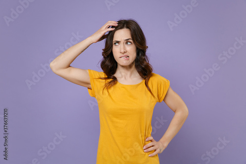 Preoccupied puzzled concerned young brunette woman 20s wearing basic yellow t-shirt posing put hand on head biting lips looking aside isolated on pastel violet colour background  studio portrait.