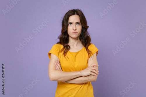 Displeased dissatisfied concerned young brunette woman 20s wearing basic yellow t-shirt posing stand holding hands crossed looking camera isolated on pastel violet colour background, studio portrait.