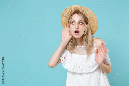 Shocked curious young blonde woman 20s wearing white summer dress hat standing try to hear you overhear listening intently looking aside isolated on blue turquoise colour background studio portrait. © ViDi Studio