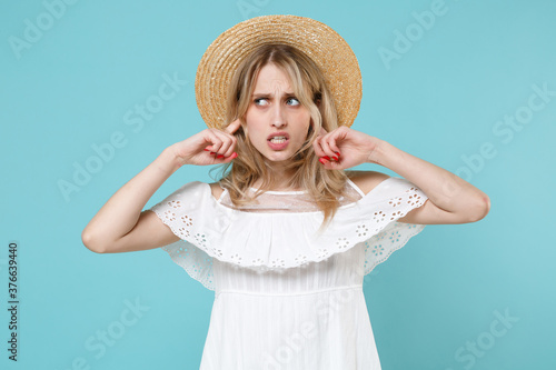 Concerned puzzled displeased confused young blonde woman 20s in white summer dress hat standing covering ears with fingers looking aside isolated on blue turquoise colour background studio portrait. © ViDi Studio