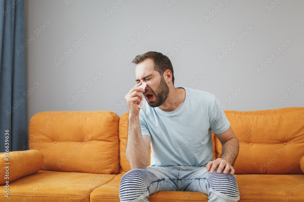 Sick exhausted young bearded man wearing casual blue t-shirt having runny nose, blowing nose to paper napkin coughing sneezing sitting on couch resting spending time in living room at home. Sick exhausted young bearded man wearing casual blue t-shirt having runny nose, blowing nose to paper napkin coughing sneezing sitting on couch resting spending time in living room at home.