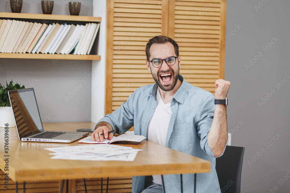 Happy joyful successful young bearded business man in blue shirt glasses sitting at desk with papers document working on laptop pc computer clenching fists doing winner gesture at home or office. Happy joyful successful young bearded business man in blue shirt glasses sitting at desk with papers document working on laptop pc computer clenching fists doing winner gesture at home or office.