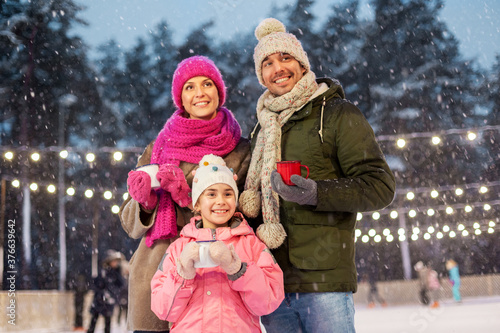 christmas, family and leisure concept - happy mother, father and daughter drinking hot tea at outdoor skating rink in winter