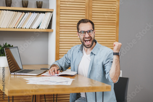 Happy joyful successful young bearded business man in blue shirt glasses sitting at desk with papers document working on laptop pc computer clenching fists doing winner gesture at home or office.