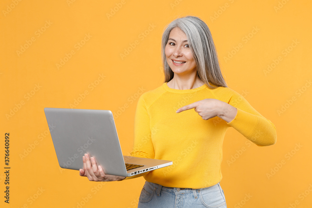 Smiling cheerful funny gray-haired asian woman wearing casual clothes standing pointing index finger on laptop pc computer looking camera isolated on bright yellow colour background studio portrait. Smiling cheerful funny gray-haired asian woman wearing casual clothes standing pointing index finger on laptop pc computer looking camera isolated on bright yellow colour background studio portrait.